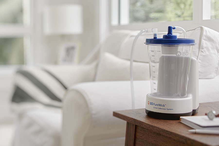 The PureWick™ Urine Collection System with battery on side table in a home setting The PureWick™ Urine Collection System with battery on side table in a home setting